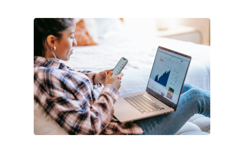 Woman checking social media engagement on her phone while using a laptop, managing side hustles from home.